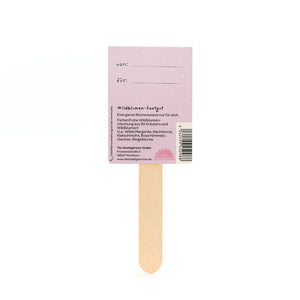 Parte posterior de un paquete de semillas de flores silvestres con un palo de madera y una etiqueta violeta que muestra los campos "de" y "para" y una descripción de la mezcla de flores silvestres.