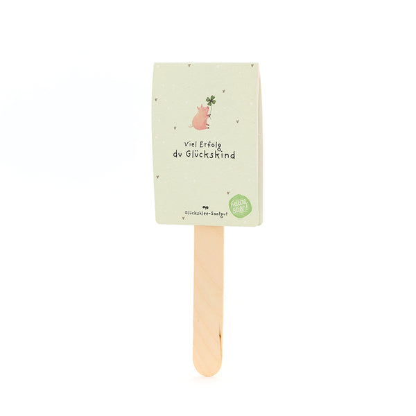 Un palo de madera con una tarjeta que muestra un cerdo con una hoja de trébol y el texto “Buena suerte, niño afortunado”.