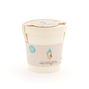Taza beige con tapa y etiqueta que representa un búho con sombrero azul y la inscripción “Hermoso cumpleañero”.
