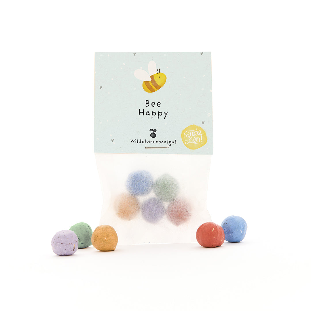 Bolsa con bombas de semillas de colores y etiqueta "Bee Happy" y una abeja dibujada.