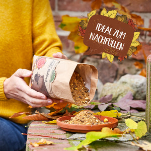 Una persona con un suéter amarillo vierte comida para erizos desde una bolsa de papel a un recipiente poco profundo rodeado de hojas de otoño; al lado hay un cartel que dice "Ideal para rellenar".