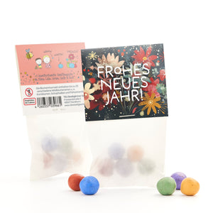 Dos bolsas de bolas de semillas de colores, una con un estampado floral que dice "¡Feliz Año Nuevo!" y otra con descripciones y dibujos, con algunas bolas en el frente.