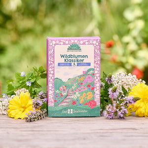 Un paquete de semillas “Wildflower Classic” con un diseño floral colorido se encuentra sobre una mesa de madera, rodeado de varias flores silvestres frescas y con una naturaleza verde borrosa en el fondo.