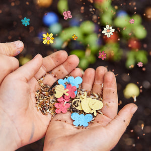 Dos manos sucias de niños sostienen semillas de flores y figuras de papel de colores con forma de flores y mariposas sobre el suelo.