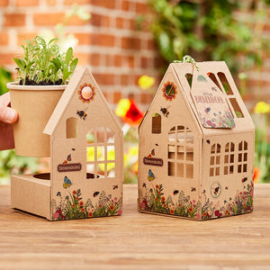 Dos casas decorativas de cartón con motivos de flores e insectos, una de ellas con una maceta que contiene plántulas verdes, ambas colocadas sobre una mesa de madera en el jardín.