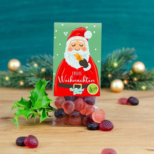 Tarjeta de felicitación navideña con una imagen de Papá Noel rodeado de hojas de acebo y gomitas de frutas, sobre una mesa de madera contra un fondo de ramas de abeto borrosas.