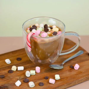 Chocolate caliente en taza transparente con malvaviscos, chispas de chocolate y bastones de caramelo, servido en una tabla de madera.