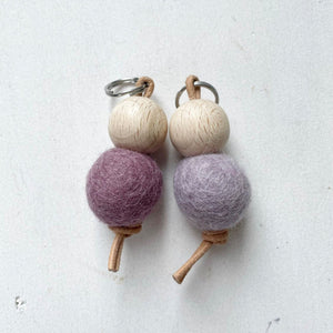 Dos llaveros hechos a mano con bolas de madera y bolas de fieltro de lana de color morado sobre fondo blanco.