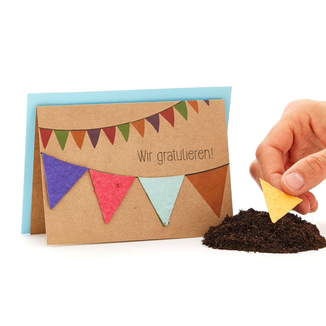 Tarjeta de felicitación con banderines de colores y la inscripción "¡Felicitaciones!", mano sosteniendo un triángulo amarillo sobre un montículo de tierra.
