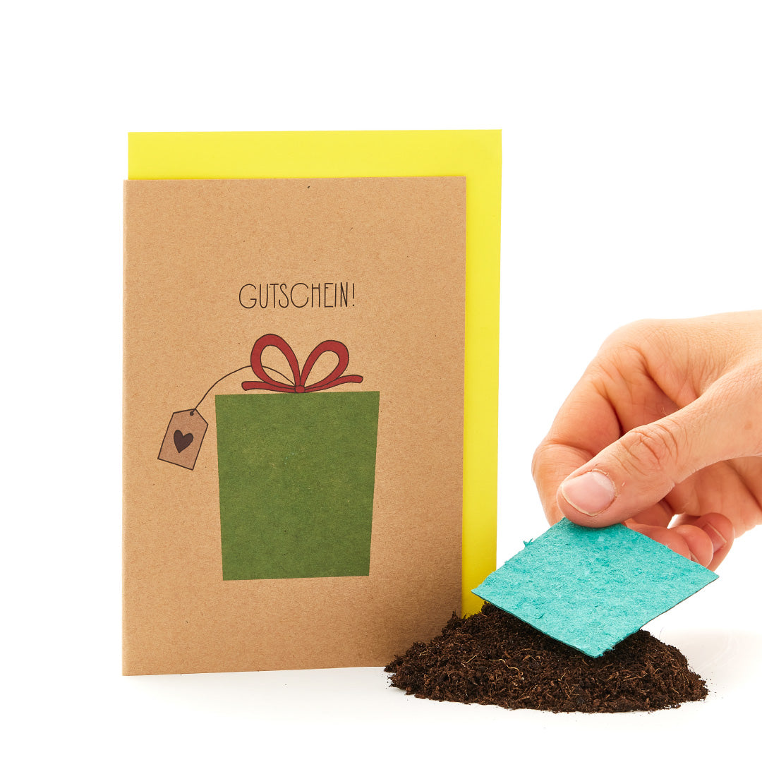 Mano sosteniendo una tarjeta de felicitación con la inscripción "VOUCHER" y un regalo dibujado, junto a un montón de tierra con un trozo de cartón azul.