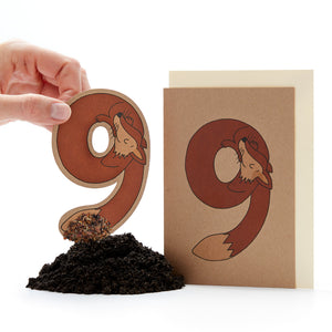 Mano sosteniendo una letra "ö" troquelada con un diseño de zorro junto a una tarjeta con el mismo diseño y un pequeño montón de tierra.