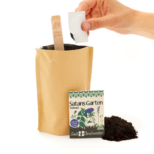 Una mano sosteniendo una bolsa de semillas sobre una bolsa de papel con tierra, junto a un cartel que dice "El jardín de Satanás" y otra bolsa de semillas.