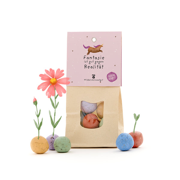 Bolsa de papel con bolas de semillas de flores silvestres de colores, algunas con pequeñas flores, y una etiqueta con un unicornio y el texto "La imaginación es buena contra la realidad".