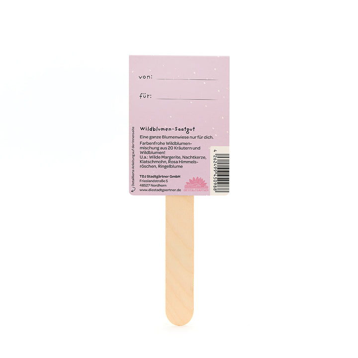 Parte posterior de un paquete de semillas de flores silvestres con un palo de madera y una etiqueta violeta que muestra los campos "de" y "para" y una descripción de la mezcla de flores silvestres.