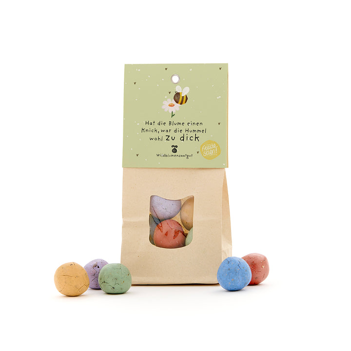 Una bolsa de papel marrón con ventana, llena de bolas de semillas de colores y una etiqueta verde con una ilustración de abeja y un dicho alemán.