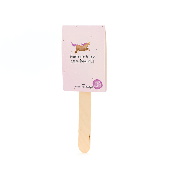 Palo de madera con una tarjeta rosa, sobre él un unicornio dibujado y el texto "La imaginación es buena contra la realidad", debajo del cual dice "Semillas de flores silvestres".
