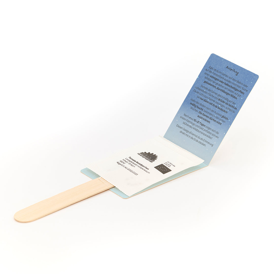 Palito de helado de madera con paquete de semillas adjunto e instrucciones desplegadas en alemán.