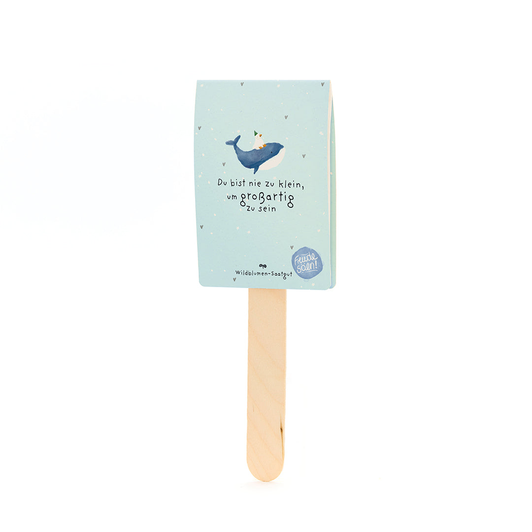 Palitos de madera con una etiqueta de papel azul claro que representa una ballena con un sombrero de fiesta y el texto "Nunca eres demasiado pequeño para ser grande".