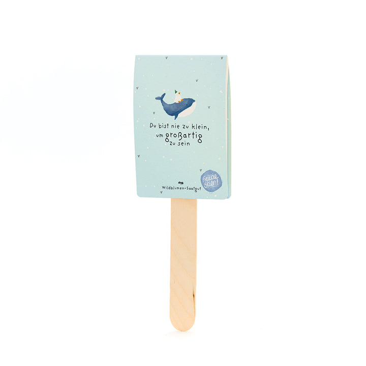 Palitos de madera con una etiqueta de papel azul claro que representa una ballena con un sombrero de fiesta y el texto "Nunca eres demasiado pequeño para ser grande".
