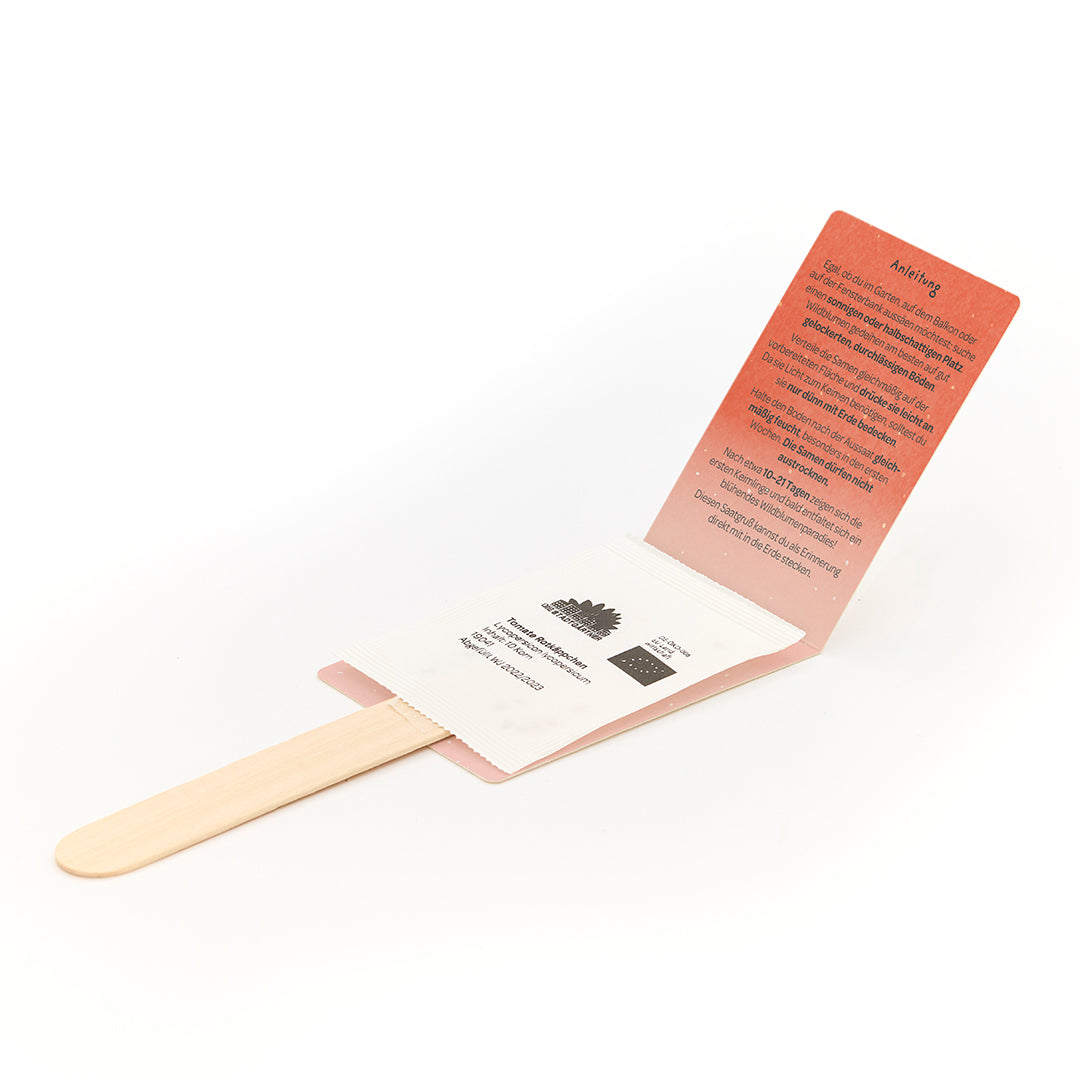 Palo de madera con paquete de semillas adjunto e instrucciones desplegadas en alemán.