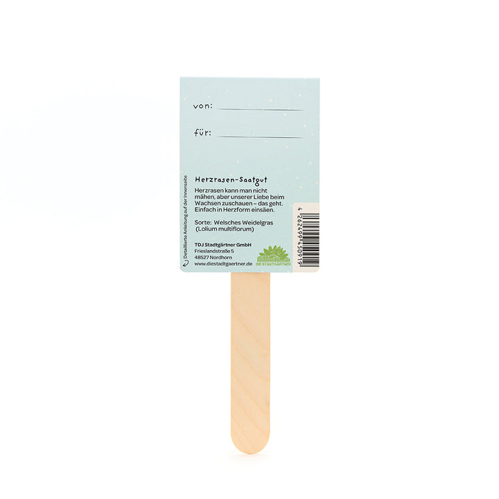 Palo de madera con una tarjeta de color azul claro adherida, con campos de texto "de" y "para", así como información sobre semillas para palpitaciones del corazón en alemán.