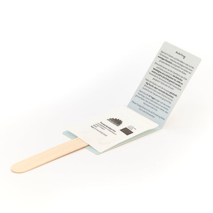 Un palo de madera con un paquete abierto que contiene una bolsa de semillas e instrucciones impresas en alemán.