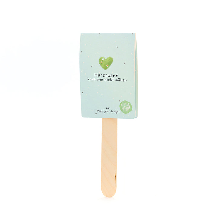 Palo de madera con un cartel de color azul claro que lleva un corazón verde y el texto "No puedes cortar un corazón que corre" y "Semillas de pasto".