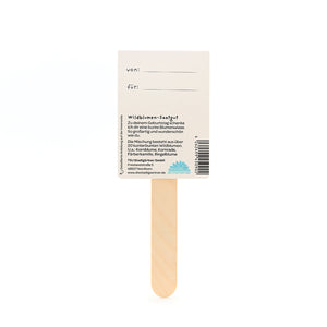 Reverso de un regalo de semillas de flores silvestres con un palo de madera, campos para "de" y "para" y una descripción de la mezcla de semillas de flores en alemán.