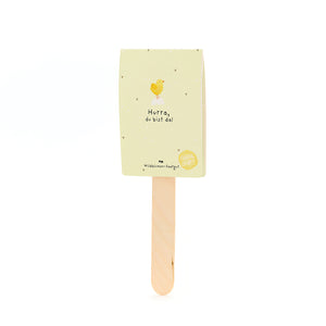 Tarjeta amarilla con ilustración de pollito y el texto "¡Hurra, estás aquí!" en un palo de madera.