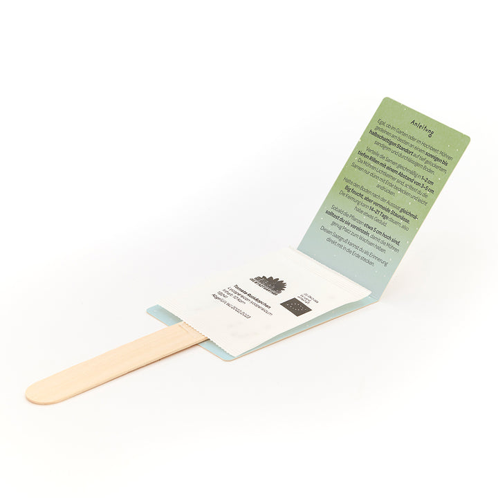 Un juego plegable con un palo de madera y un paquete de semillas, así como una tarjeta de instrucciones verde en alemán.