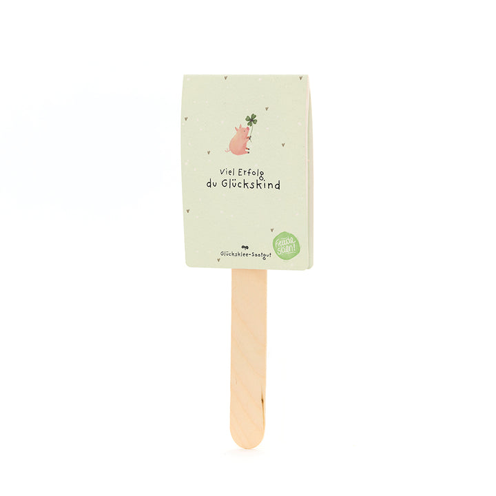 Un palo de madera con una tarjeta que muestra un cerdo con una hoja de trébol y el texto “Buena suerte, niño afortunado”.