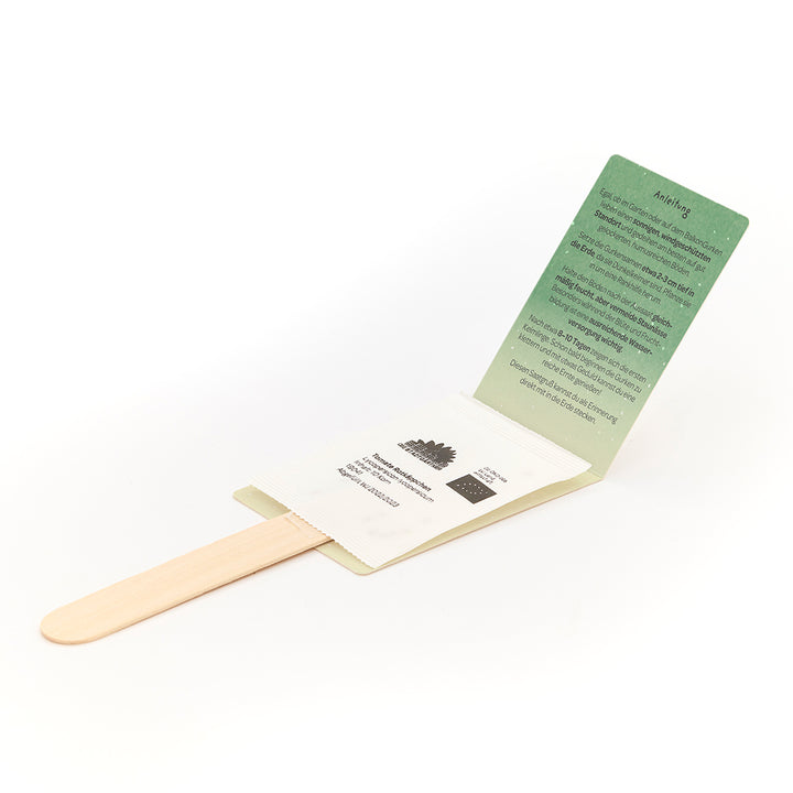 Palo de madera con sobre adjunto que contiene una tarjeta de instrucciones verde y un paquete de semillas blancas.