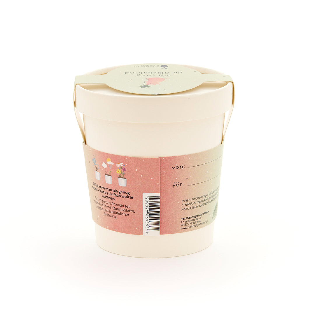 Maceta beige con asa, etiqueta rosa con ilustraciones de plantas y texto en alemán, etiqueta de regalo en la tapa.