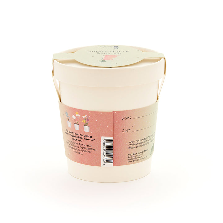 Maceta beige con asa, etiqueta rosa con ilustraciones de plantas y texto en alemán, etiqueta de regalo en la tapa.