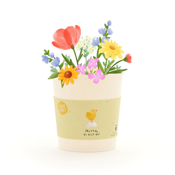 Maceta ilustrada con flores de colores y una etiqueta amarilla que representa un pollito y el texto "¡Hurra, estás aquí!".