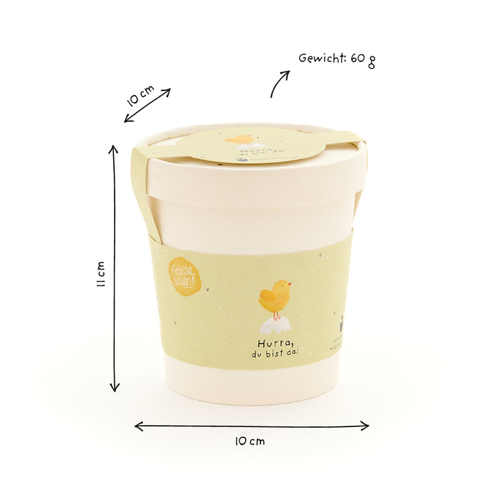 Taza beige con etiqueta amarilla, ilustración de pollito y texto "¡Hurra, estás aquí!", con dimensiones (10 cm de diámetro, 11 cm de altura) y peso (60 g) en alemán.