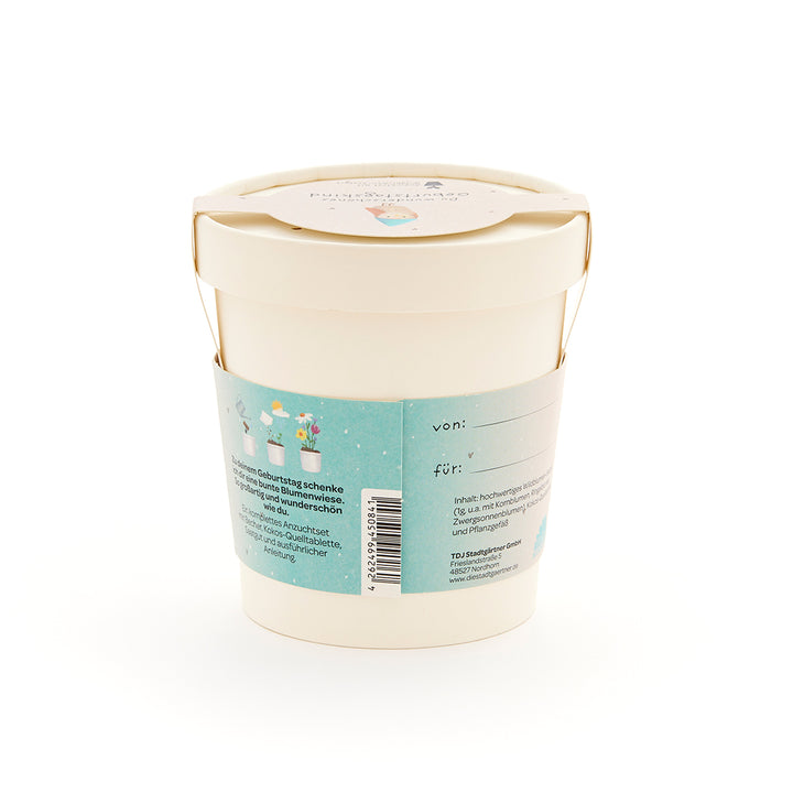 Maceta beige con etiqueta y asa azules, juego de regalo para un prado de flores, etiquetas "de" y "para" visibles.