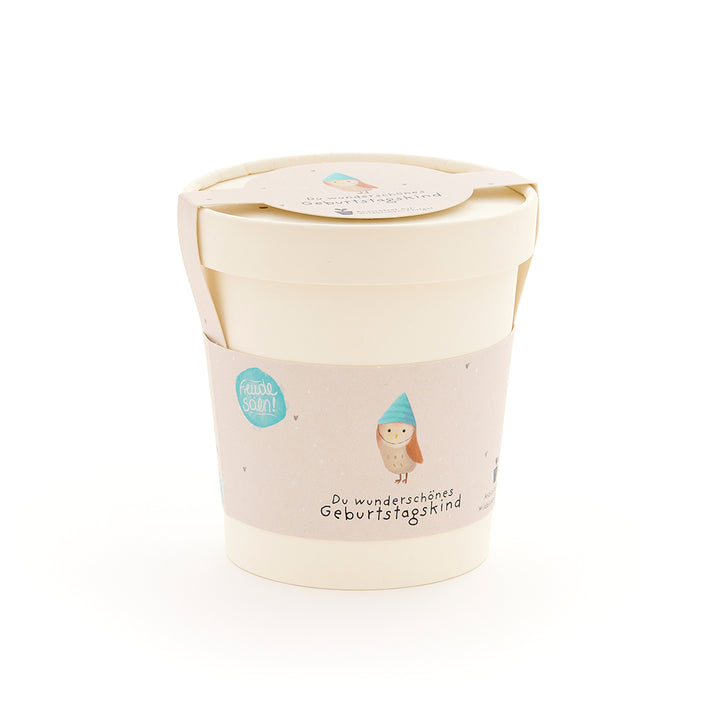 Taza beige con tapa y etiqueta que representa un búho con sombrero azul y la inscripción “Hermoso cumpleañero”.