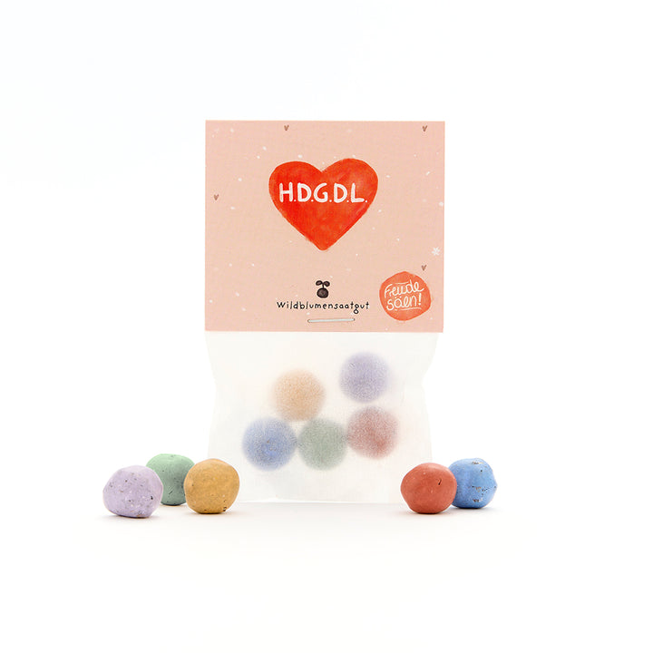 Bolsa de bolas de semillas de colores, el empaque dice "H.D.G.D.L" en un corazón rojo y "semillas de flores silvestres".