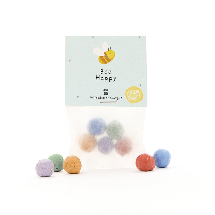 Bolsa con bombas de semillas de colores y etiqueta "Bee Happy" y una abeja dibujada.