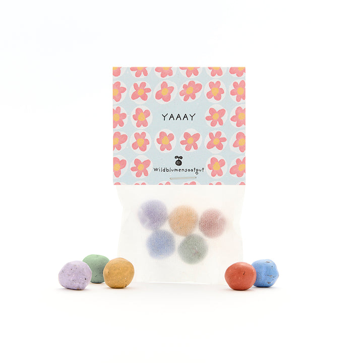Bolas de semillas multicolores en un paquete transparente con una etiqueta floral y la inscripción "YAAAY".