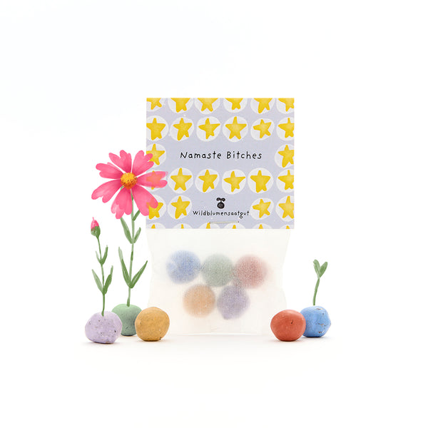 Bolsa de bombas de semillas de colores con la inscripción "Namaste Bitches", junto a flores silvestres en flor y germinando sobre un fondo blanco.