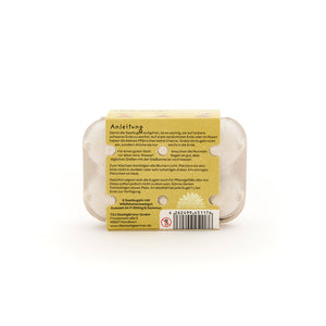 Parte posterior de un pequeño cartón de huevos con instrucciones adjuntas para sembrar semillas, código de barras e información del fabricante en alemán.