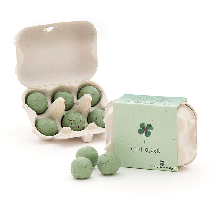 Seis bolas de semillas verdes en un pequeño cartón de huevos, junto a una caja cerrada con una hoja de trébol y las palabras "Buena suerte".
