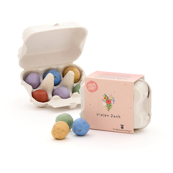 Seis bombas de semillas de colores en un pequeño paquete de cartón de huevos, junto a una caja cerrada con la inscripción "Gracias" y una ilustración de flores.