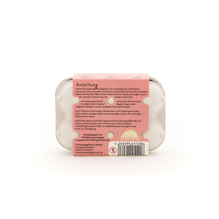Una pequeña caja de cartón con una etiqueta de instrucciones rosa y bolas de semillas, con instrucciones de cuidado en alemán en la parte posterior.