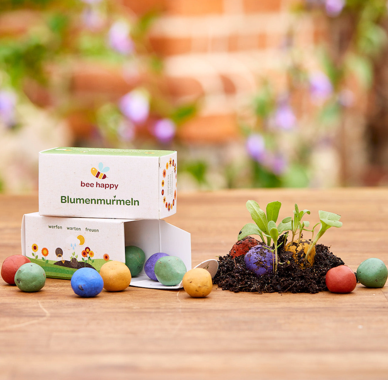 Canicas de flores de colores y una caja etiquetada "canicas de flores felices para las abejas" sobre una mesa de madera, con tierra y plantas brotando al lado.