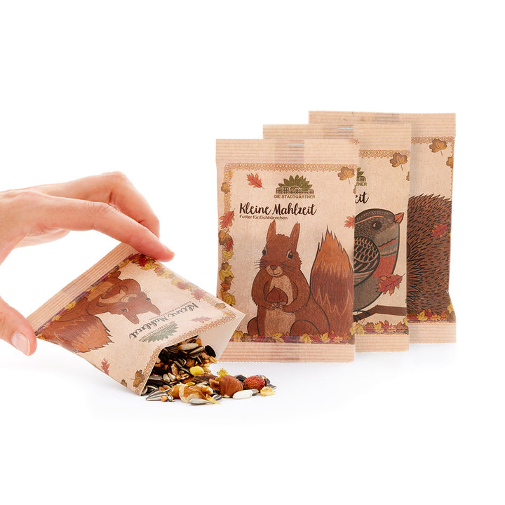Una mano abre una pequeña bolsa de papel con comida para mascotas junto a otras dos bolsas con ilustraciones de ardillas y erizos.