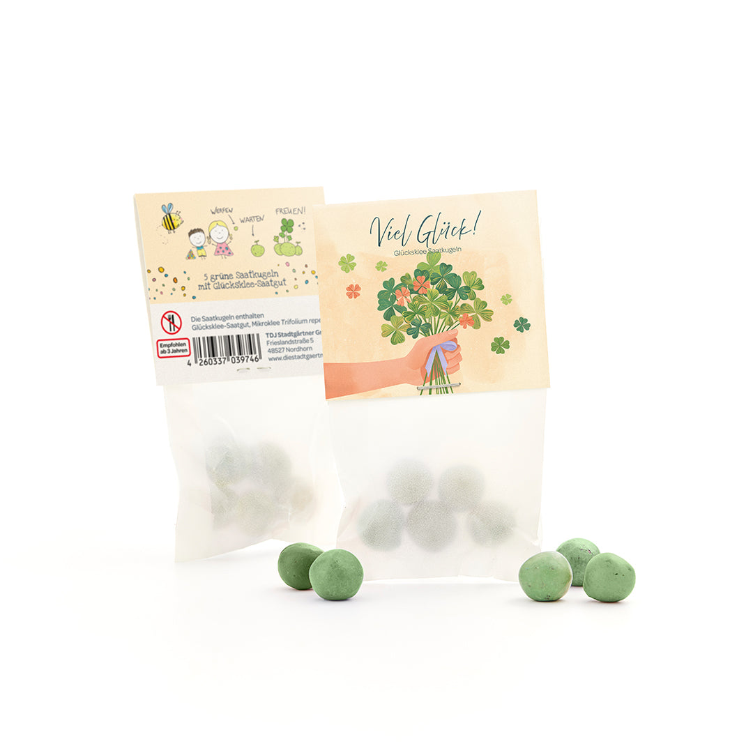 Bolsa con bolas de semillas verdes y semillas de trébol de la suerte, junto a varias bolas verdes sueltas, en el embalaje un motivo de felicitación y una ilustración de trébol.