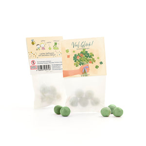 Bolsa con bolas de semillas verdes y semillas de trébol de la suerte, junto a varias bolas verdes sueltas, en el embalaje un motivo de felicitación y una ilustración de trébol.